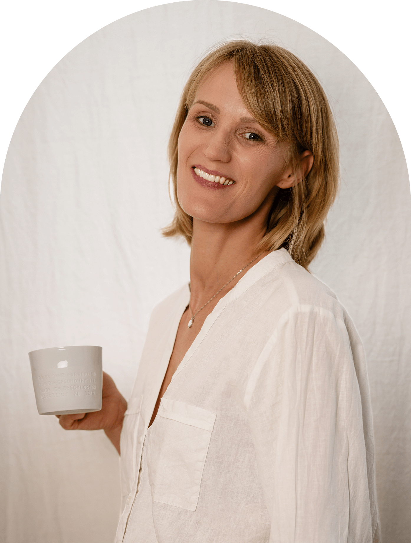 Health Coach Miri Heinrich steht freundlich lachend vor einer weißen Wand und hält einen Becher in der Hand